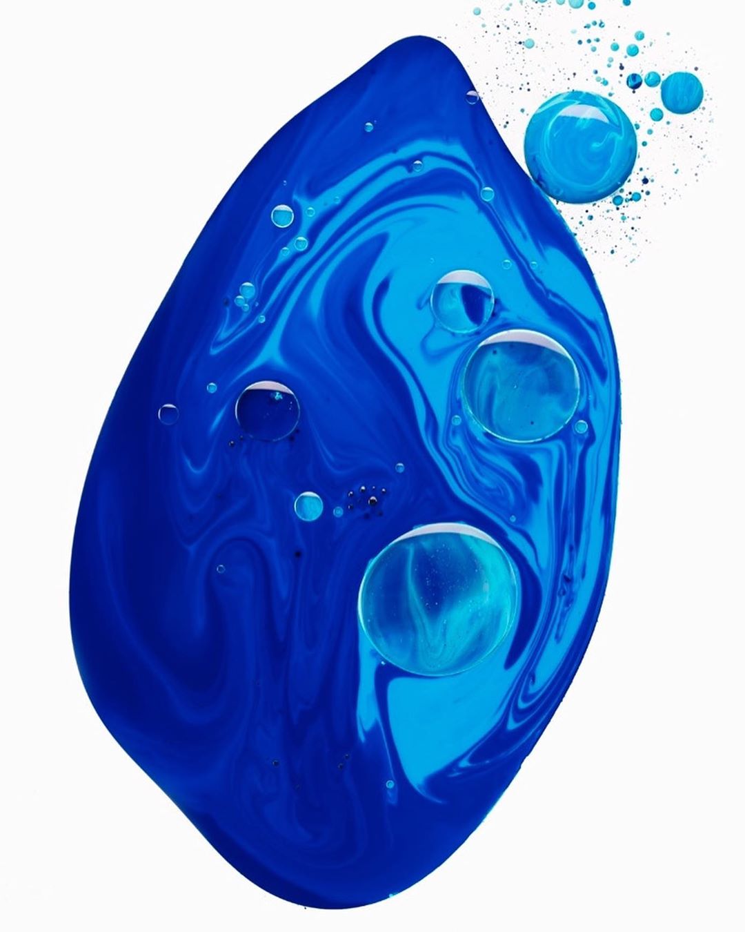 Abstract still life with dark and light blue liquid paint forming a leaf-like shape with small water droplets on a white background.