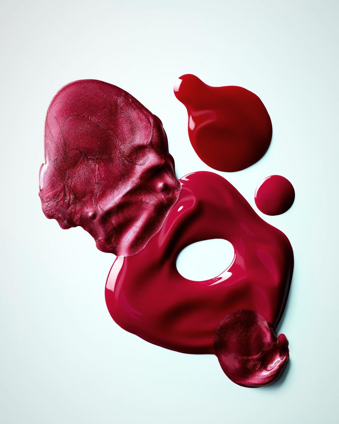 Overhead shot of various smears of red-colored nail polish on a white background. The nail polish is glossy and viscous.