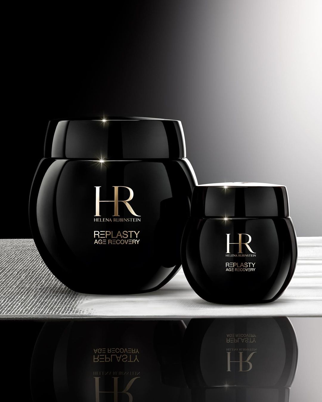 Two black Helena Rubinstein Re-Plasty Age Recovery cream jars of different sizes are displayed on a white surface with reflections.
