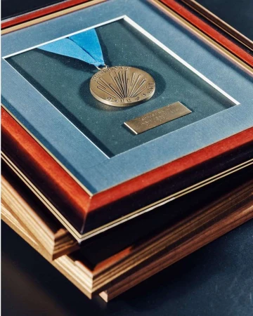 Framed medal on display with a blue ribbon, encased in a layered wooden frame against a dark backdrop, presented in portrait format.