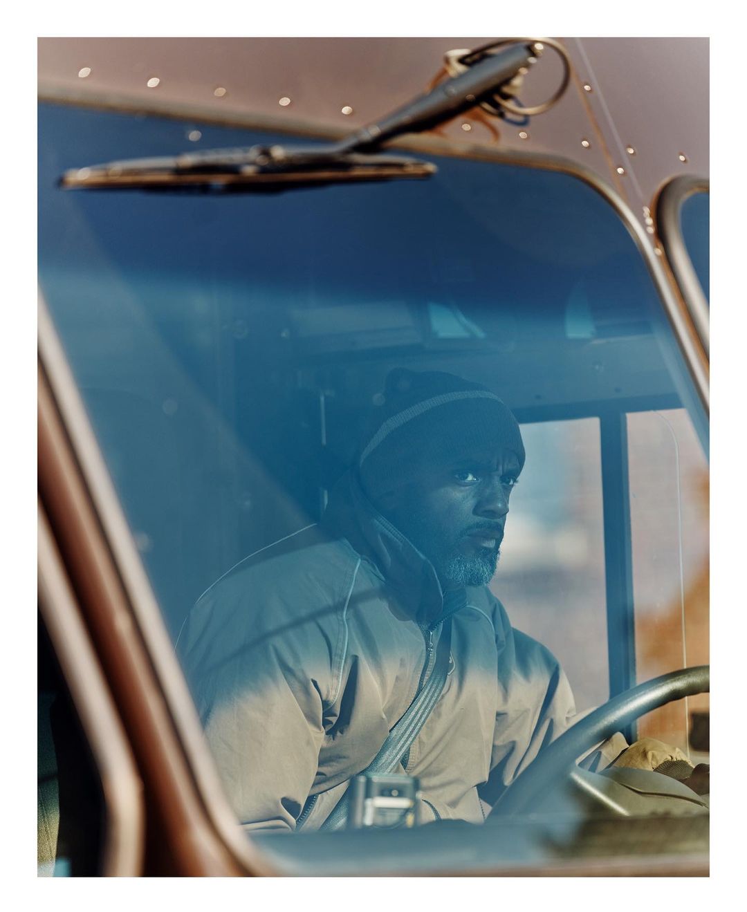 Man with beard wearing a beanie and jacket sits in the driver's seat of a truck, viewed through the blue-tinted windshield.