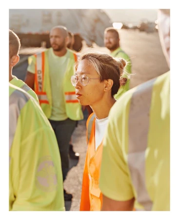 A group of construction workers wearing neon yellow and orange reflective vests are standing outdoors, looking to the left.