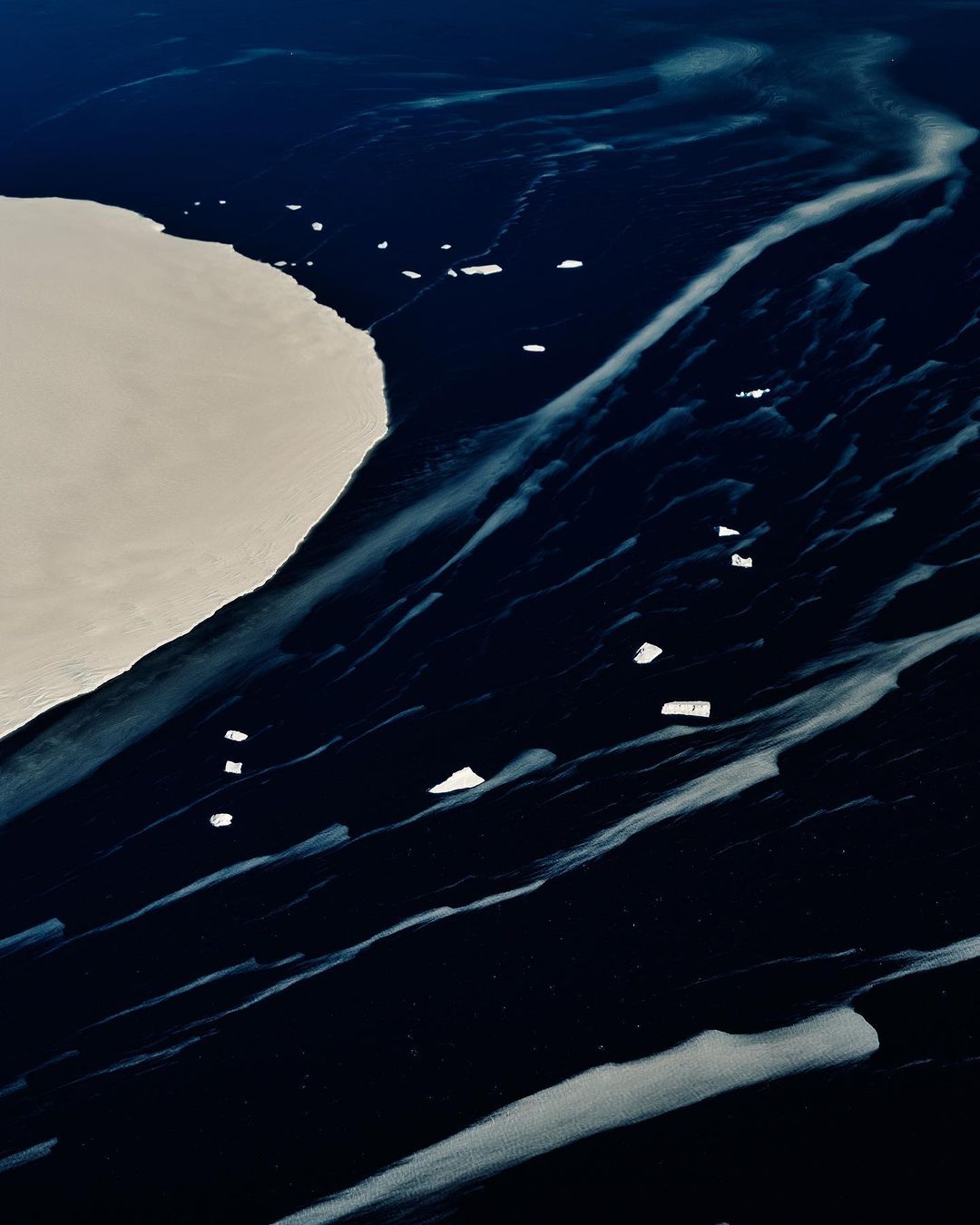 Aerial view of dark ocean water with icy formations and an iceberg, showcasing nature's textures and the meeting of water and ice.