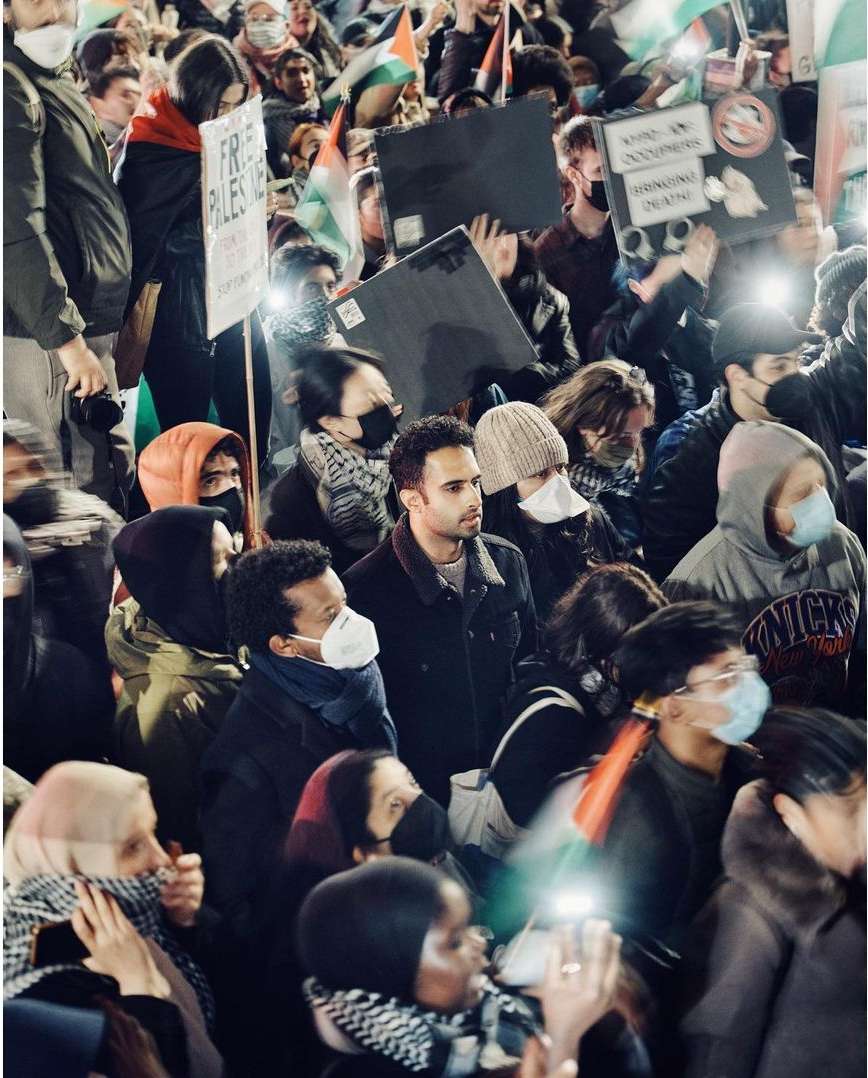 Crowd of people gather, holding signs and Palestinian flags in a night protest with a blurry motion effect for a sense of scale.
