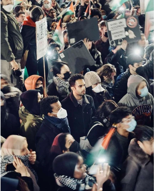 Crowd of people gather, holding signs and Palestinian flags in a night protest with a blurry motion effect for a sense of scale.
