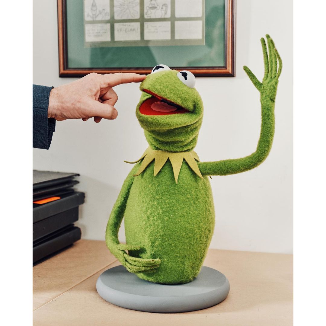 Kermit the Frog puppet on a gray stand being touched on the nose, with framed artwork hanging on the wall in the background.