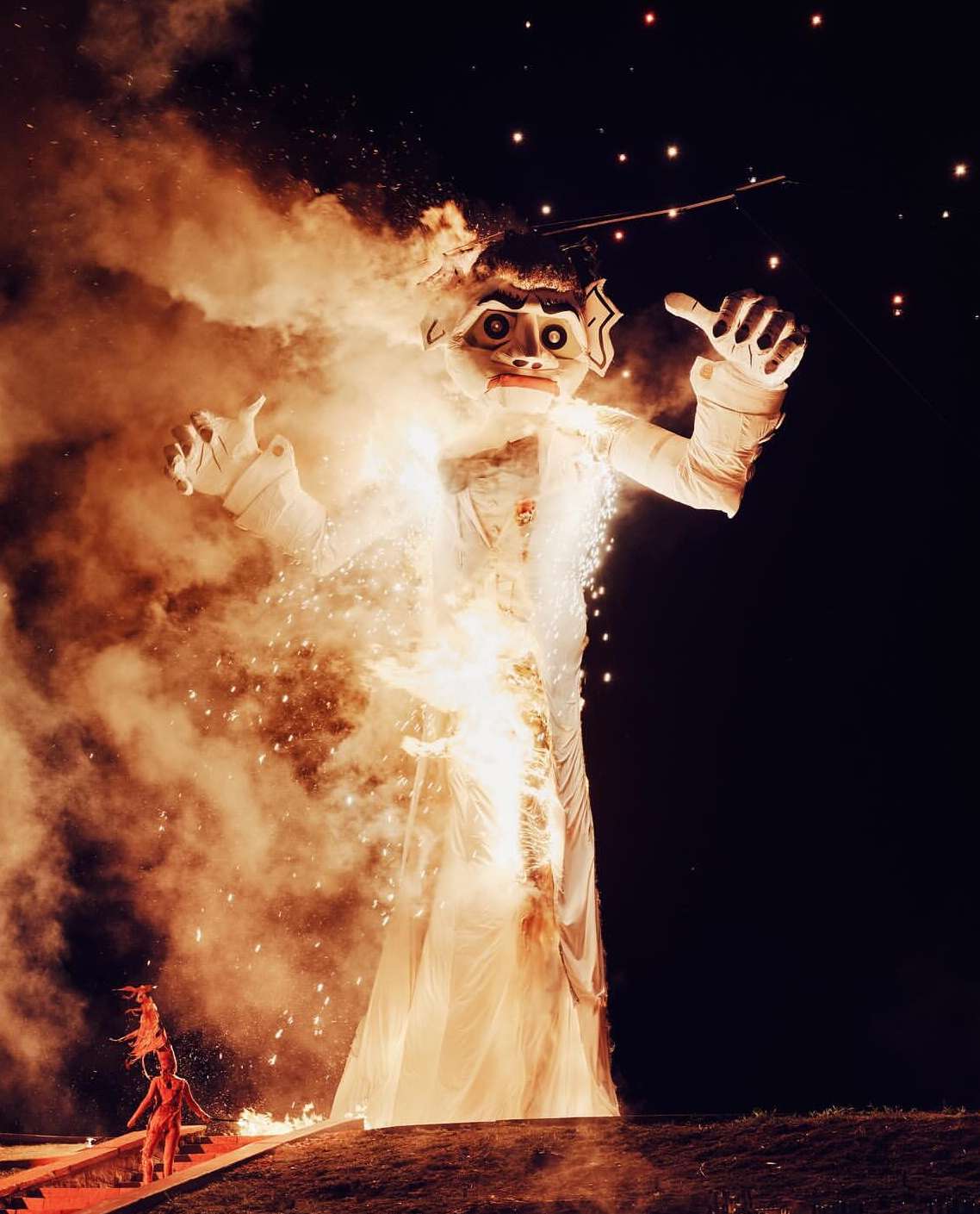 Large puppet-like figure surrounded by fire and smoke at night with small people at the base, possibly part of a performance or festival.
