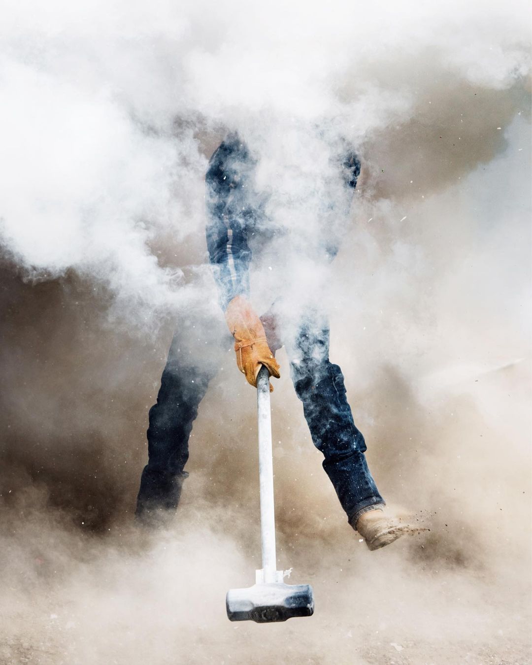 A person in jeans and work boots swings a sledgehammer, surrounded by a cloud of dust and debris against a bright background.