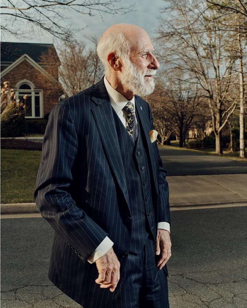 Elderly man in a pinstripe suit standing on a street near a house surrounded by trees on a bright day. Elegant attire.