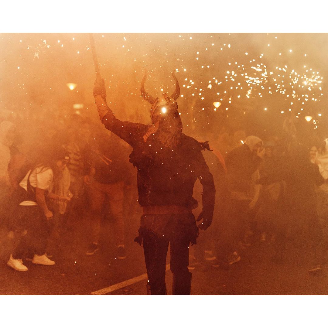 A costumed figure in a horned helmet holds a long object aloft amidst orange smoke and sparks, surrounded by a crowd during a street festival.