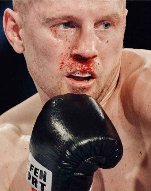 Close-up of a male boxer with blood on his face, wearing a black boxing glove, highlighting the intensity and physicality of the sport.