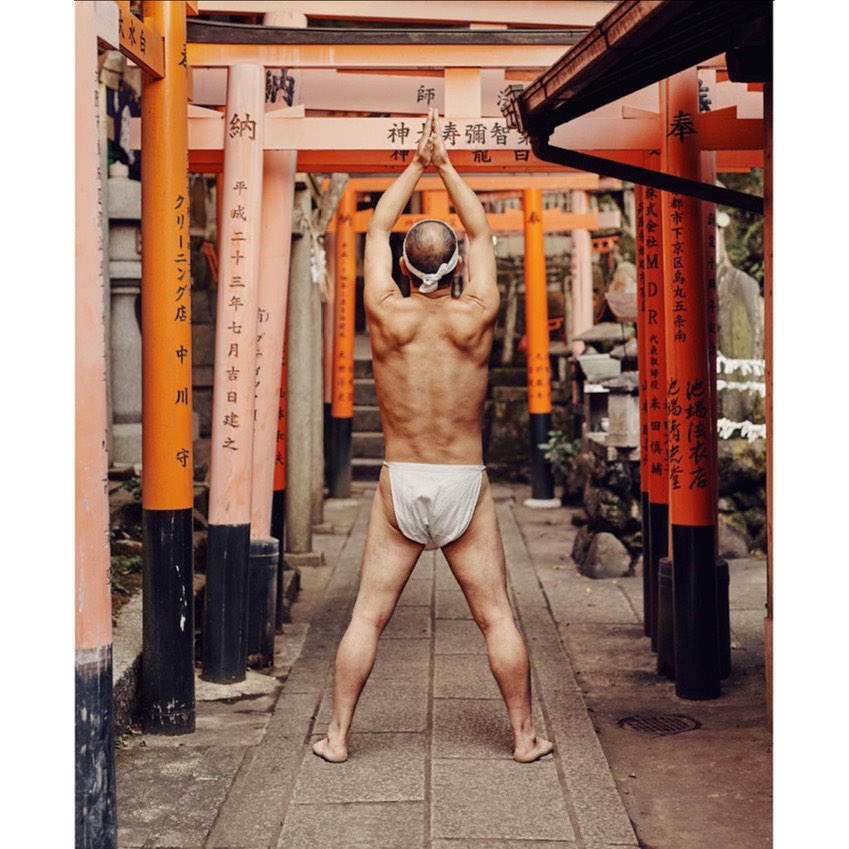 Bare-chested man in traditional Japanese dress stands among orange torii gates, arms raised in prayerful pose, outdoors on stone path.
