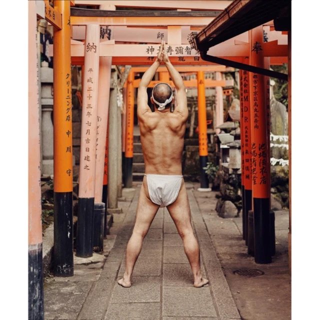 Bare-chested man in traditional Japanese dress stands among orange torii gates, arms raised in prayerful pose, outdoors on stone path.