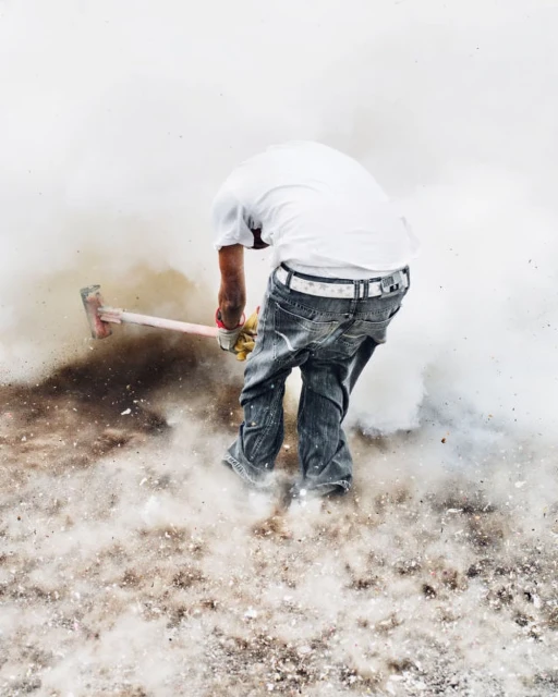Man in jeans and white shirt swings a sledgehammer against a dusty, light background, action shot.