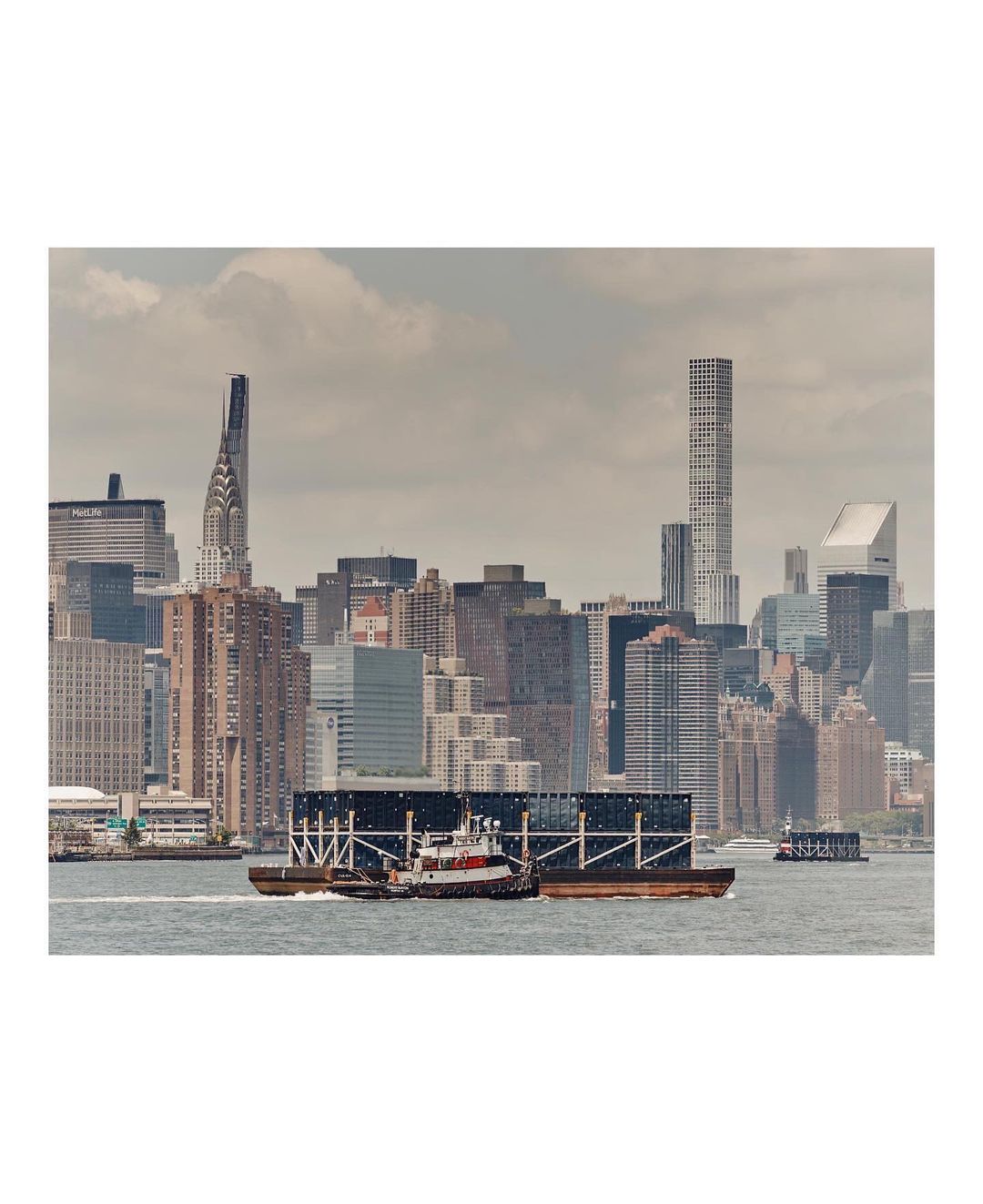 Tugboat pulls a barge on the water with a cityscape featuring the Chrysler Building and modern skyscrapers in the background, cloudy day.