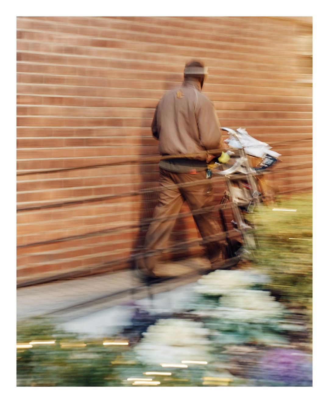 Blurred view of an adult male walking past a brick wall with a metal cart outside in daylight, creating a sense of motion and urban life.