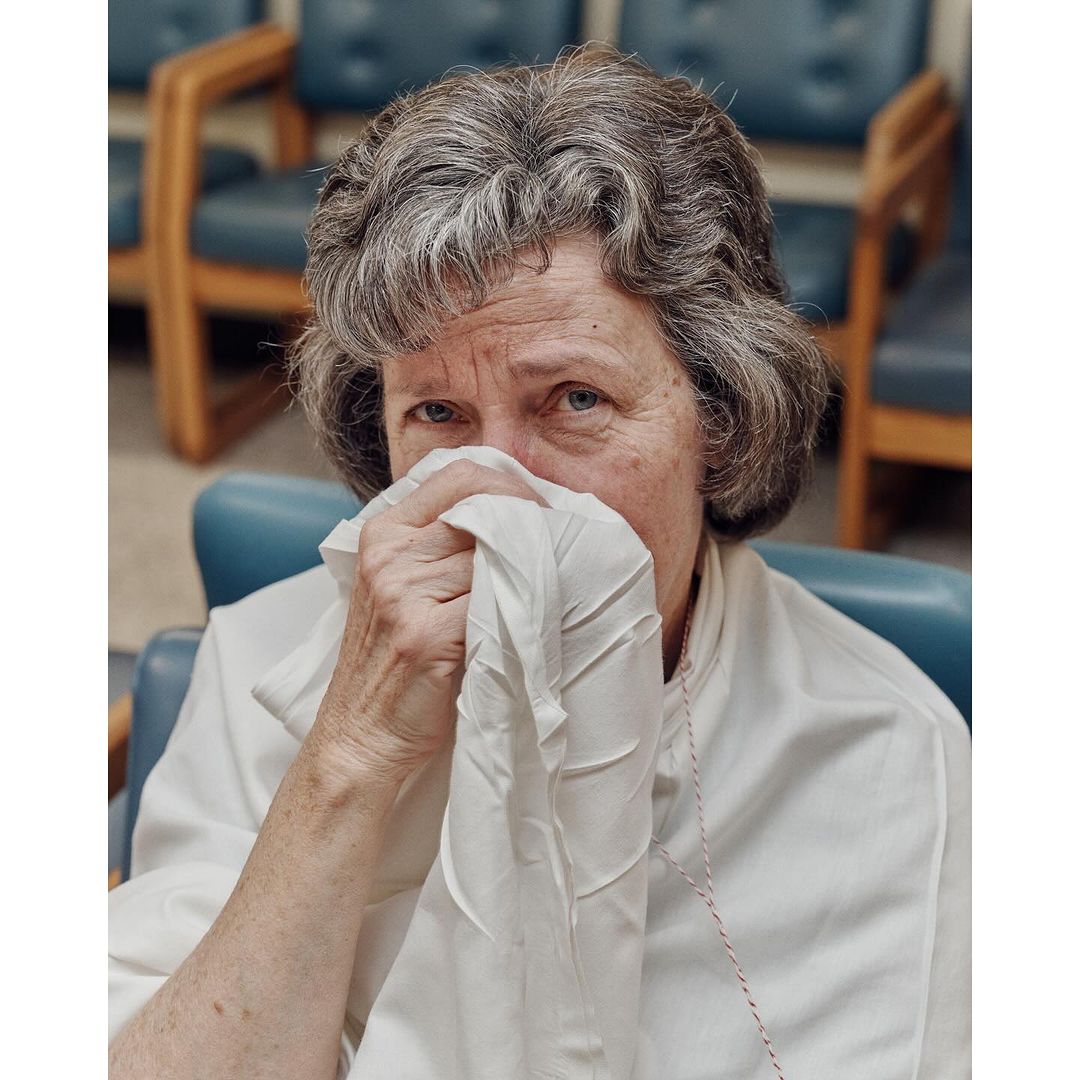 Indoor medium shot of an elderly woman with gray hair covering her face with a white tissue while sitting in a blue chair.