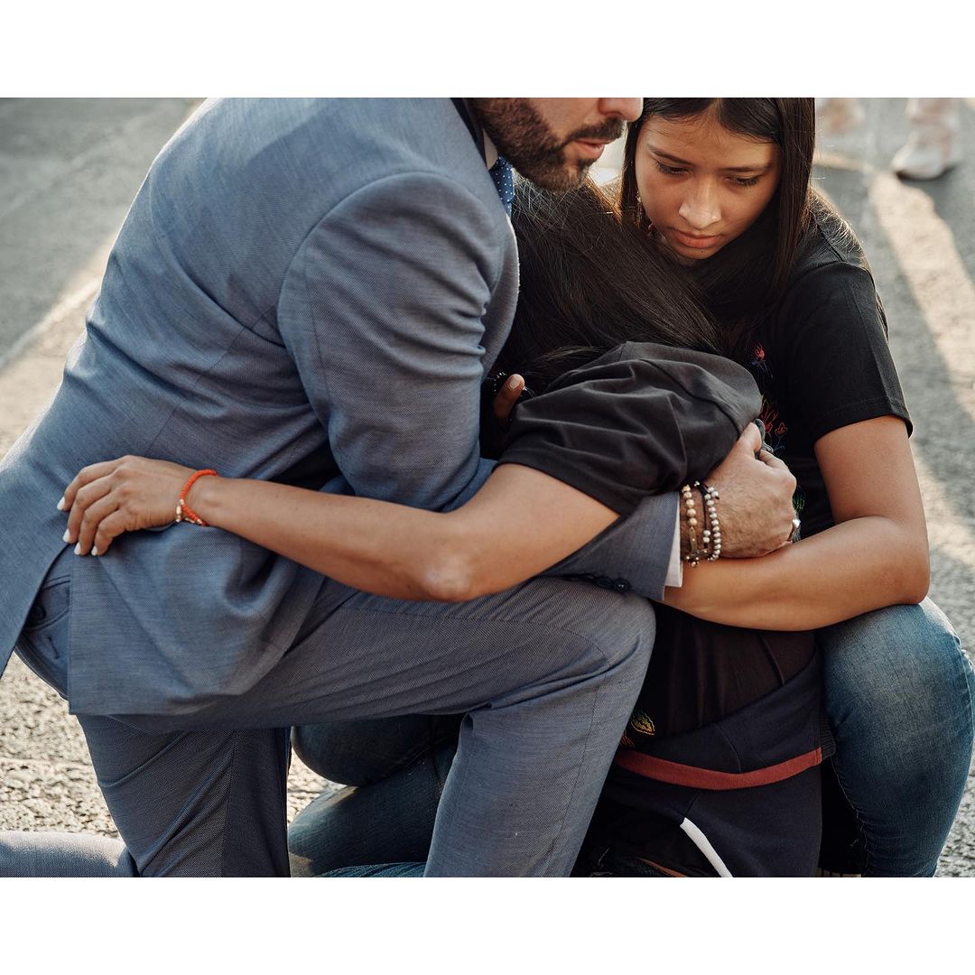 Man and young woman embrace outdoors. He wears a grey suit and she wears a black shirt. They kneel on a street.