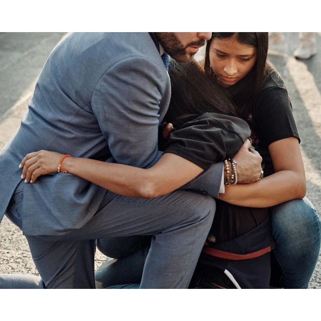 Man and young woman embrace outdoors. He wears a grey suit and she wears a black shirt. They kneel on a street.