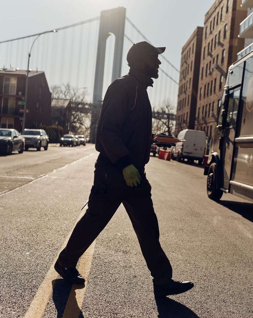 An adult man silhouetted while crossing a city street in daylight, with a large suspension bridge and buildings in the background.