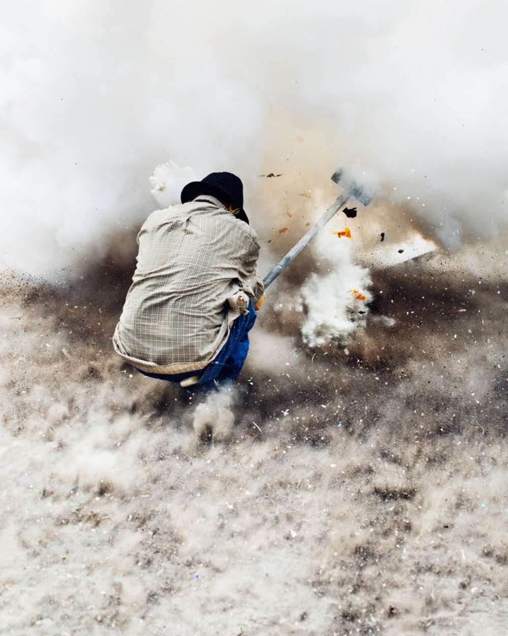 An adult working outdoors amidst a cloud of dust and debris, swinging a tool. The background shows a bright, hazy atmosphere.