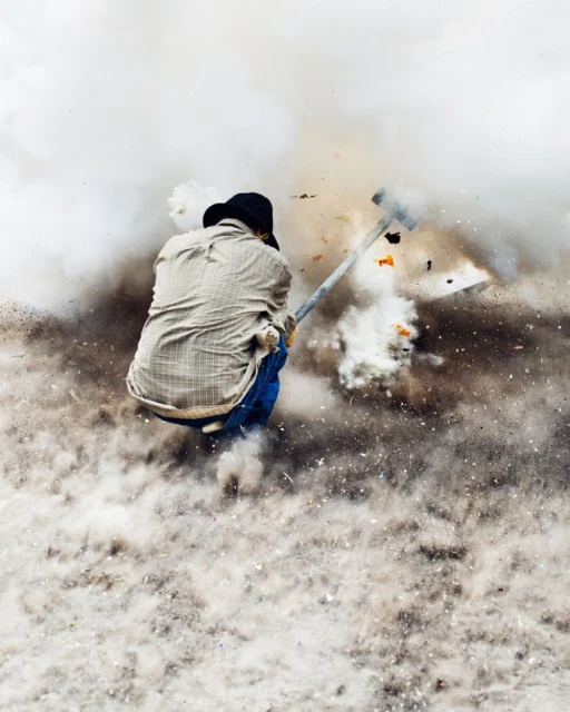 An adult working outdoors amidst a cloud of dust and debris, swinging a tool. The background shows a bright, hazy atmosphere.