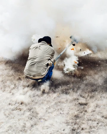An adult working outdoors amidst a cloud of dust and debris, swinging a tool. The background shows a bright, hazy atmosphere.