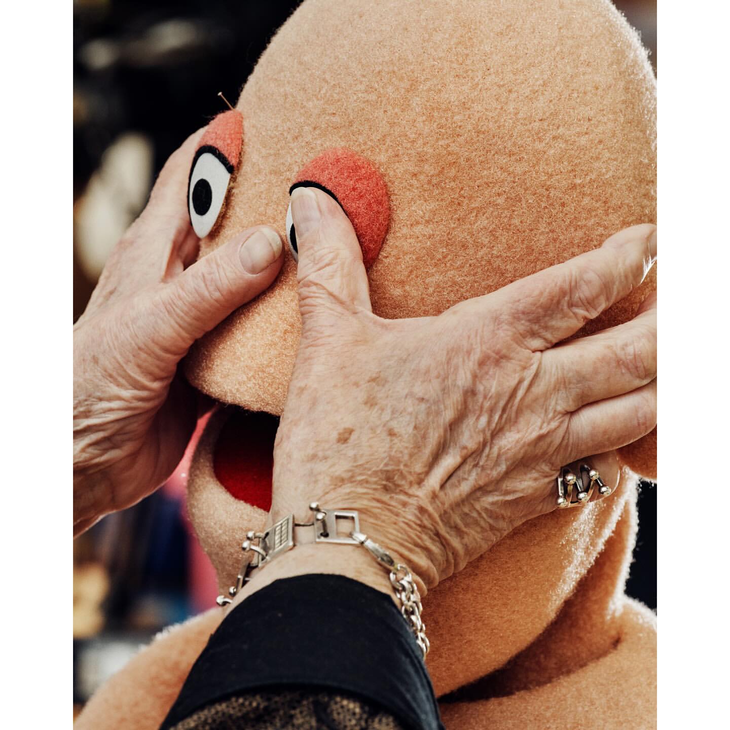Close-up of a person's hands adjusting the eye of a tan puppet head with visible seam, set against a dark, blurred background.