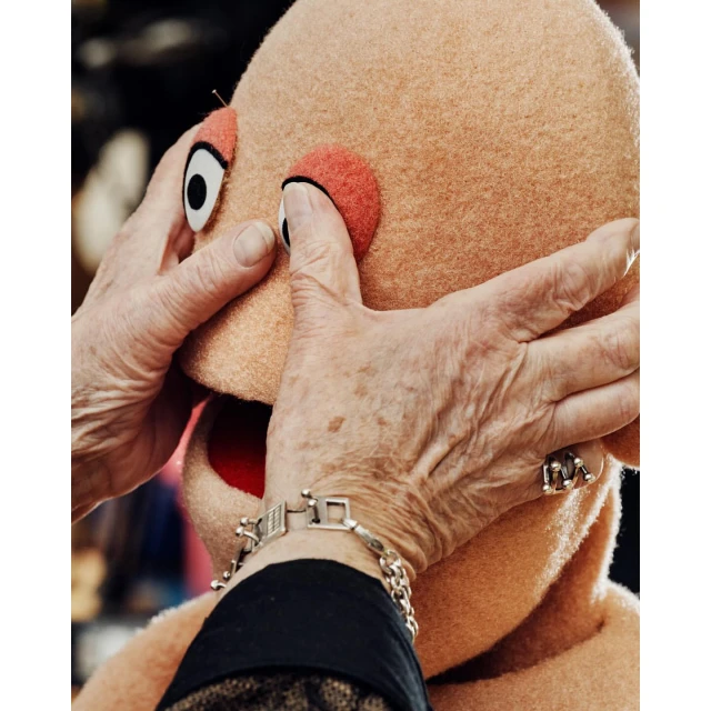 Close-up of a person's hands adjusting the eye of a tan puppet head with visible seam, set against a dark, blurred background.