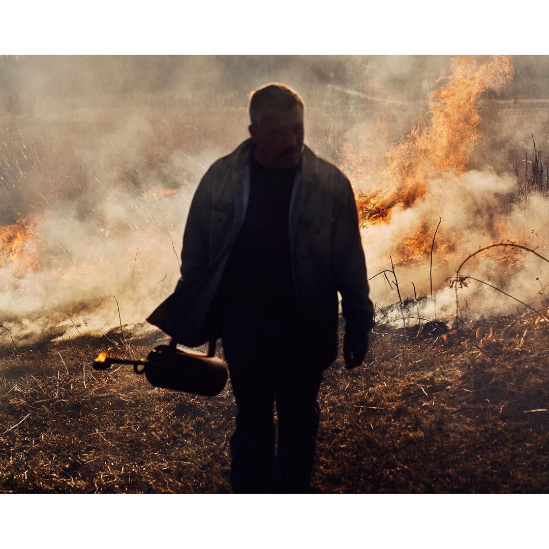 Man uses a flamethrower in a field with a fire burning in the background, wearing gloves and denim shirt in a rural environment.