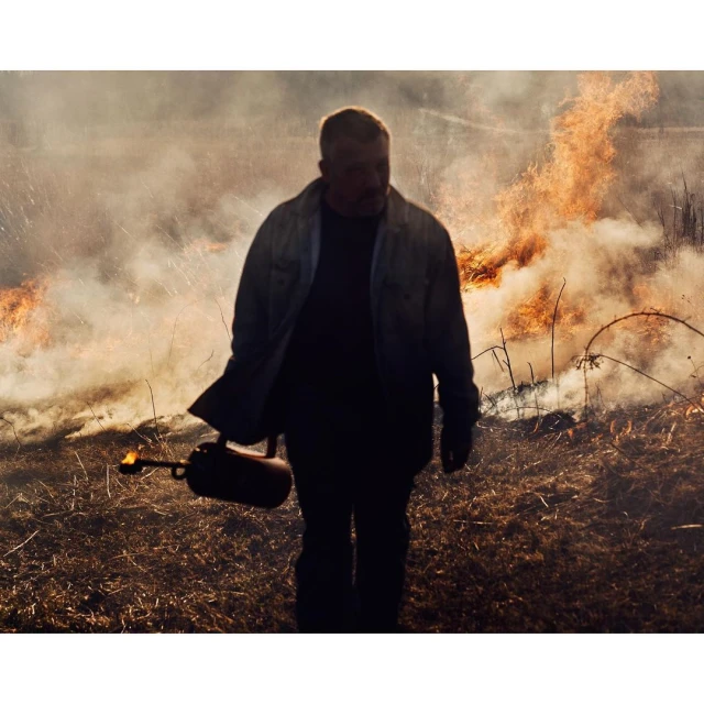 Man uses a flamethrower in a field with a fire burning in the background, wearing gloves and denim shirt in a rural environment.