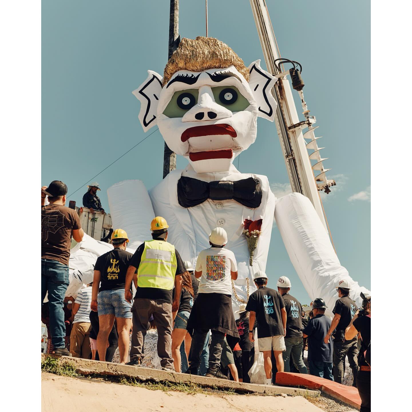 Large puppet figure of a man in a suit is suspended by a crane as a group of people on the ground look up at it against a blue sky.