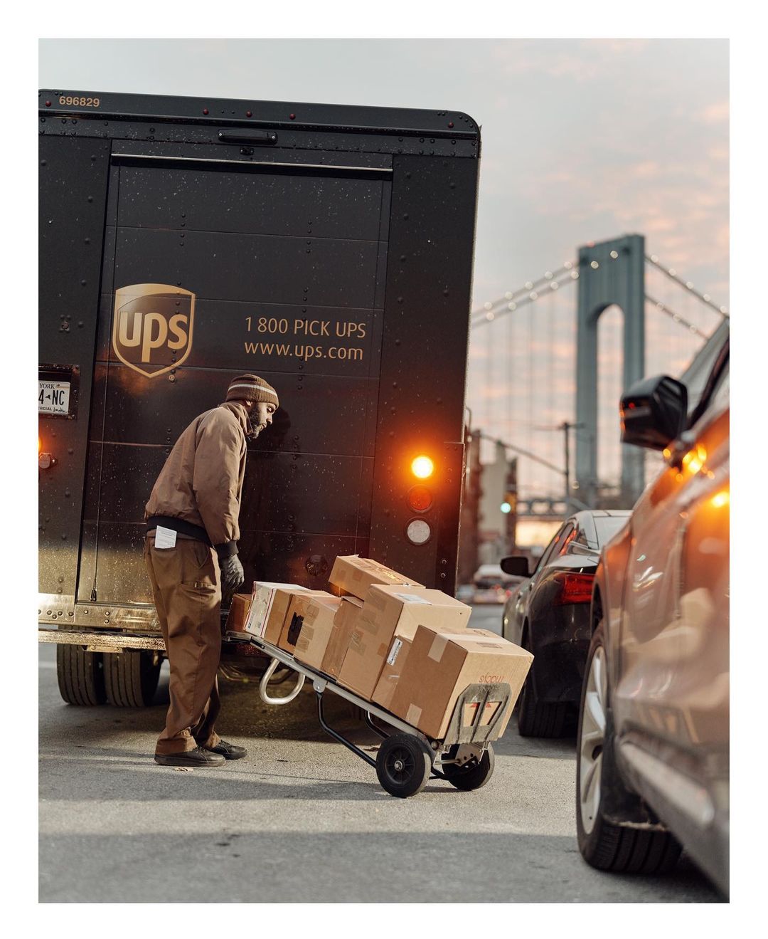 UPS delivery person transports boxes on a dolly in an urban setting with a bridge in the background, alongside vehicles.