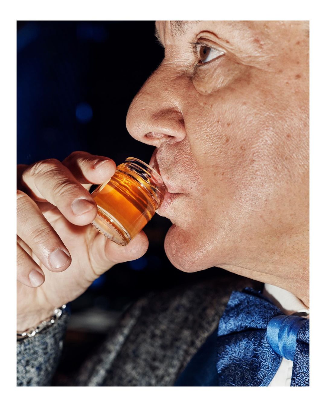 Close-up portrait of a mature man in a suit drinking from a shot glass with an amber-colored liquid, against a dark background with bokeh.
