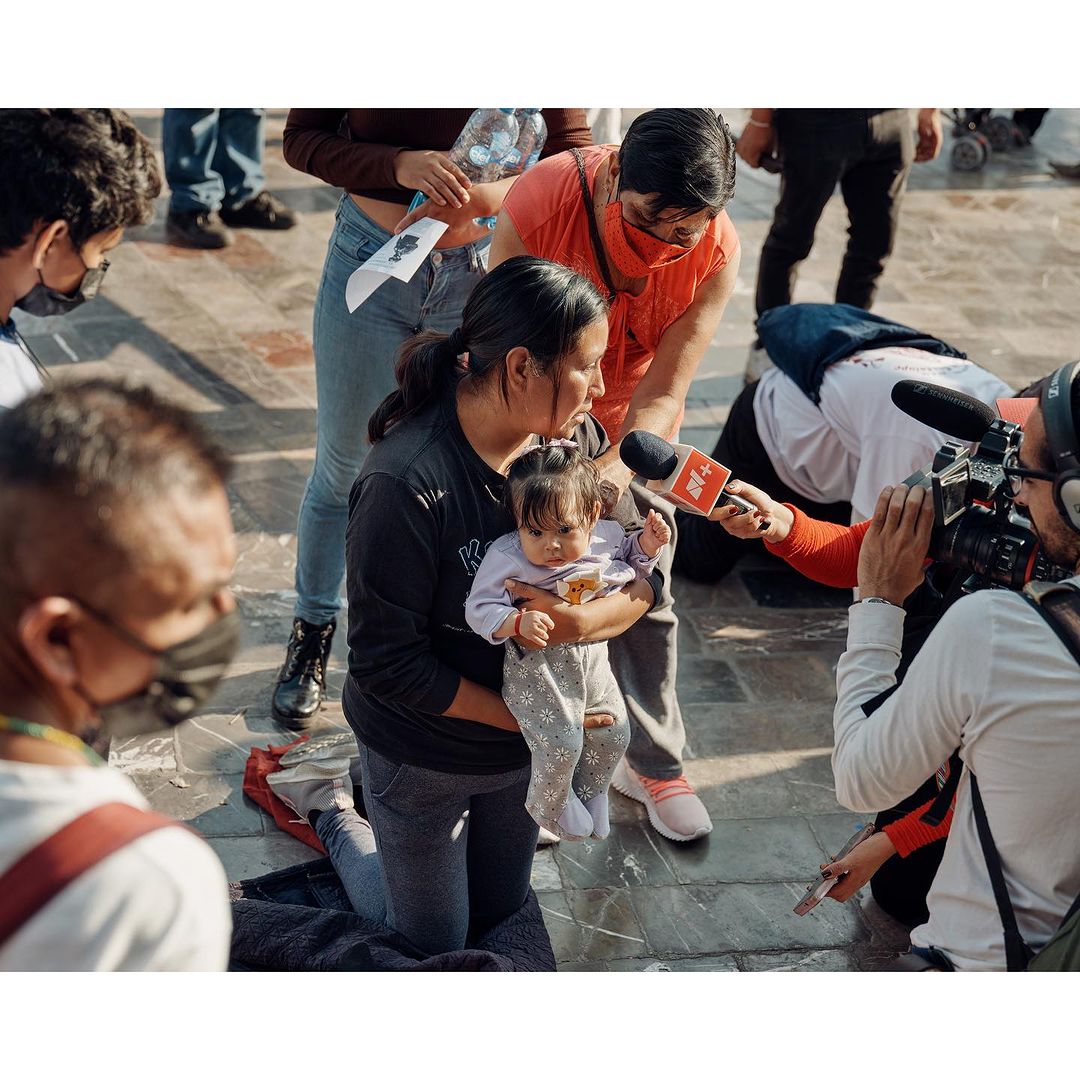 Woman holding baby at press conference with reporters and cameras surrounding them in an outdoor urban environment on a sunny day.