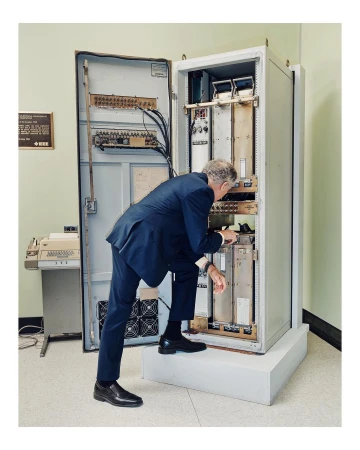 Man in a suit inspects vintage computer hardware inside an open server rack, standing on a white block in an indoor setting with an old typewriter.