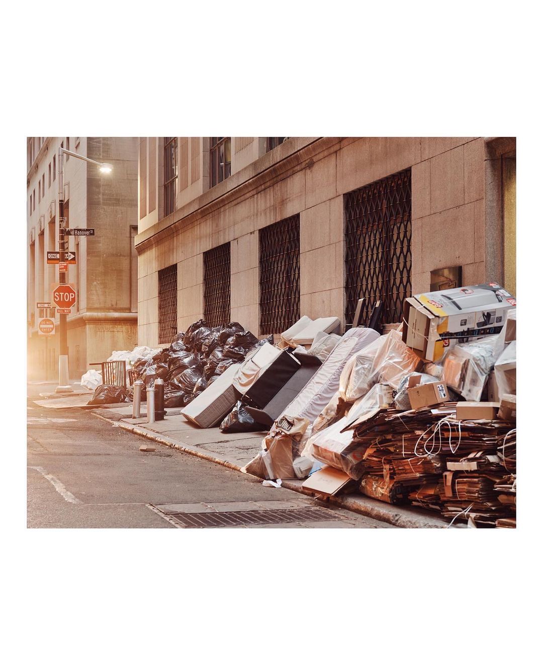 Urban street scene with piles of garbage bags and discarded items including furniture, boxes, and paper strewn along the sidewalk.