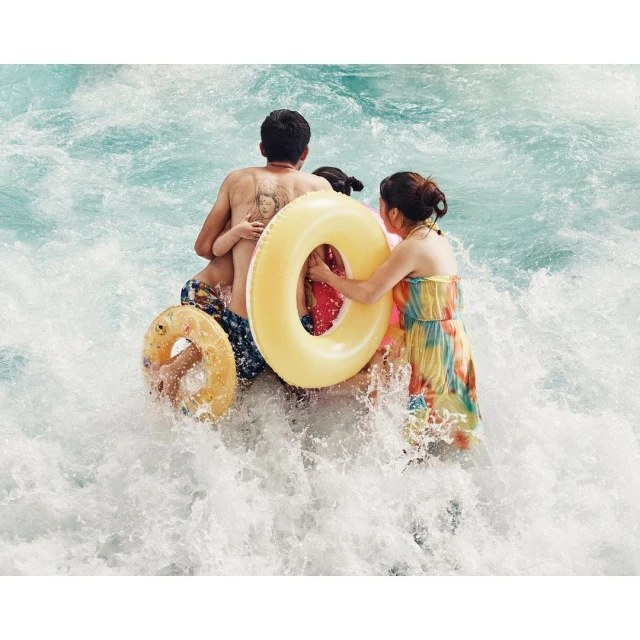 Family with two kids wading into rushing water with inflatable yellow rings. A woman in a dress follows behind them.