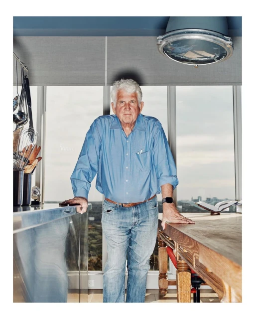 Portrait of an elderly man in a blue shirt standing in a modern kitchen with natural light from large windows.