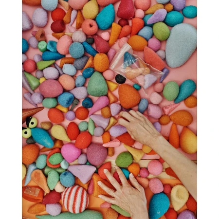 Hands amidst a colorful assortment of felted shapes, arranged on a light pink surface, capturing a vibrant and tactile composition.