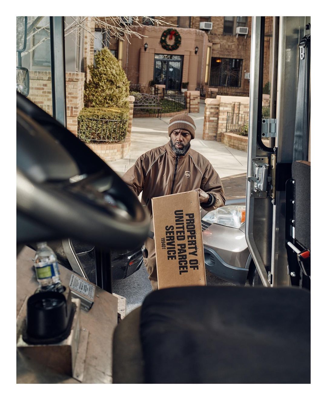 Delivery man in brown uniform carries a package from his truck to a building with a wreath, captured from inside the vehicle.