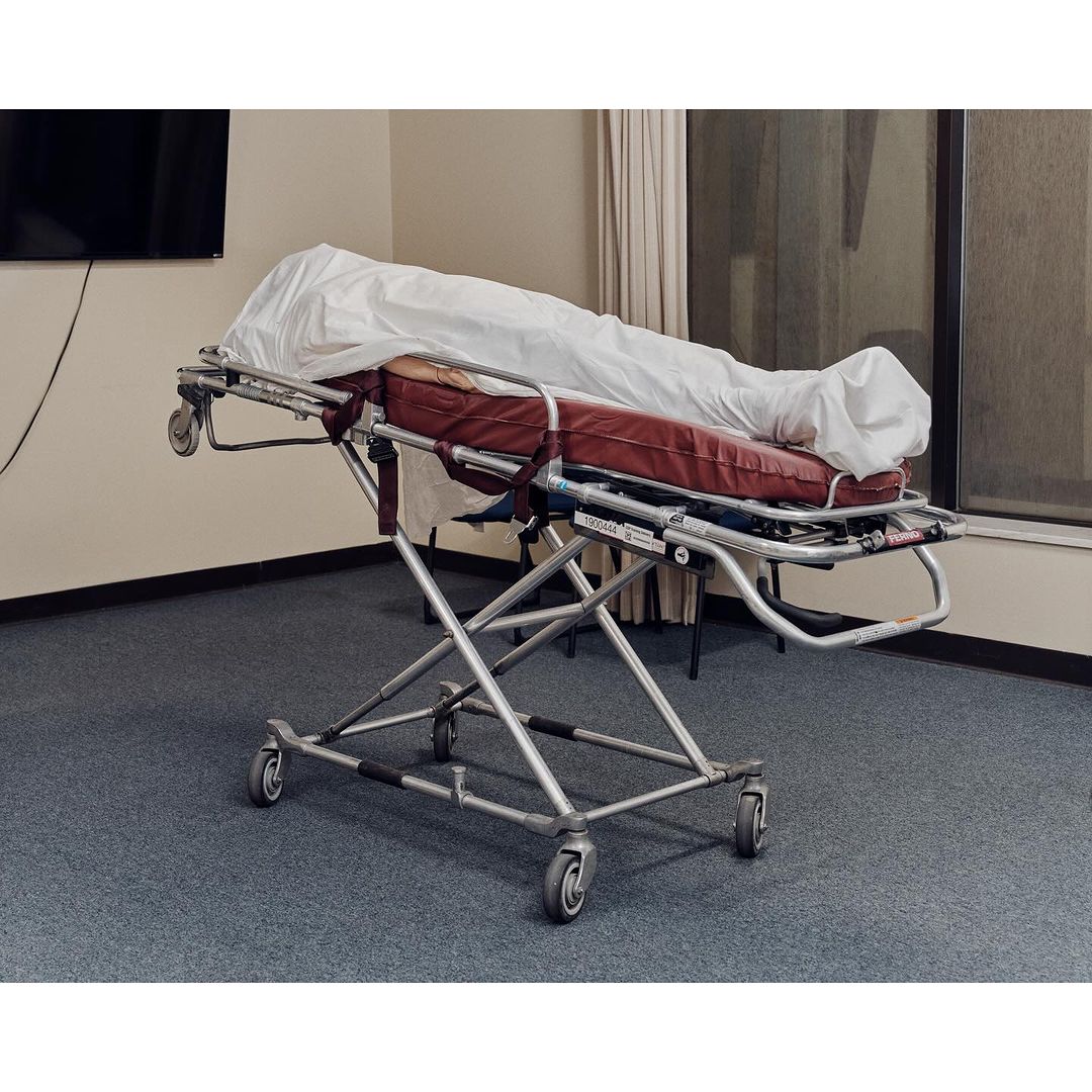 Hospital gurney with white sheet in an indoor medical environment with window and wall-mounted television, reflecting a clinical setting.