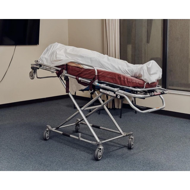 Hospital gurney with white sheet in an indoor medical environment with window and wall-mounted television, reflecting a clinical setting.