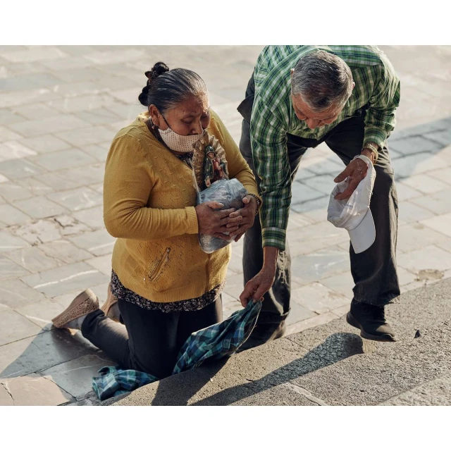 Woman kneeling with wrapped object, receiving garment from standing person in a plaid shirt. Urban outdoor setting with paving stones.