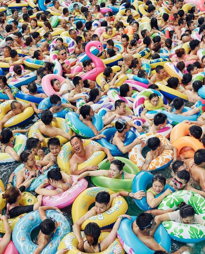 Aerial view captures a large group of people swimming in a pool with colorful inflatable tubes, creating a vibrant, crowded water scene.