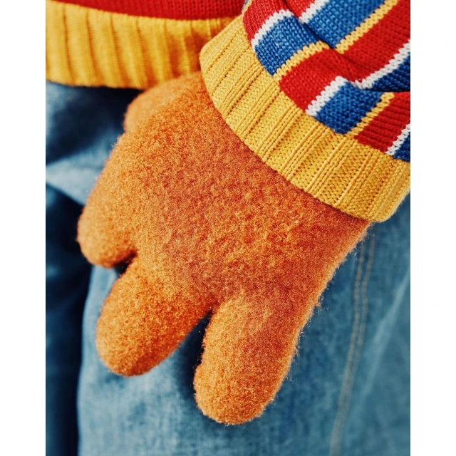 Close-up of an orange knit glove on a person wearing a striped sweater and blue jeans, capturing a warm and tactile winter fashion detail.
