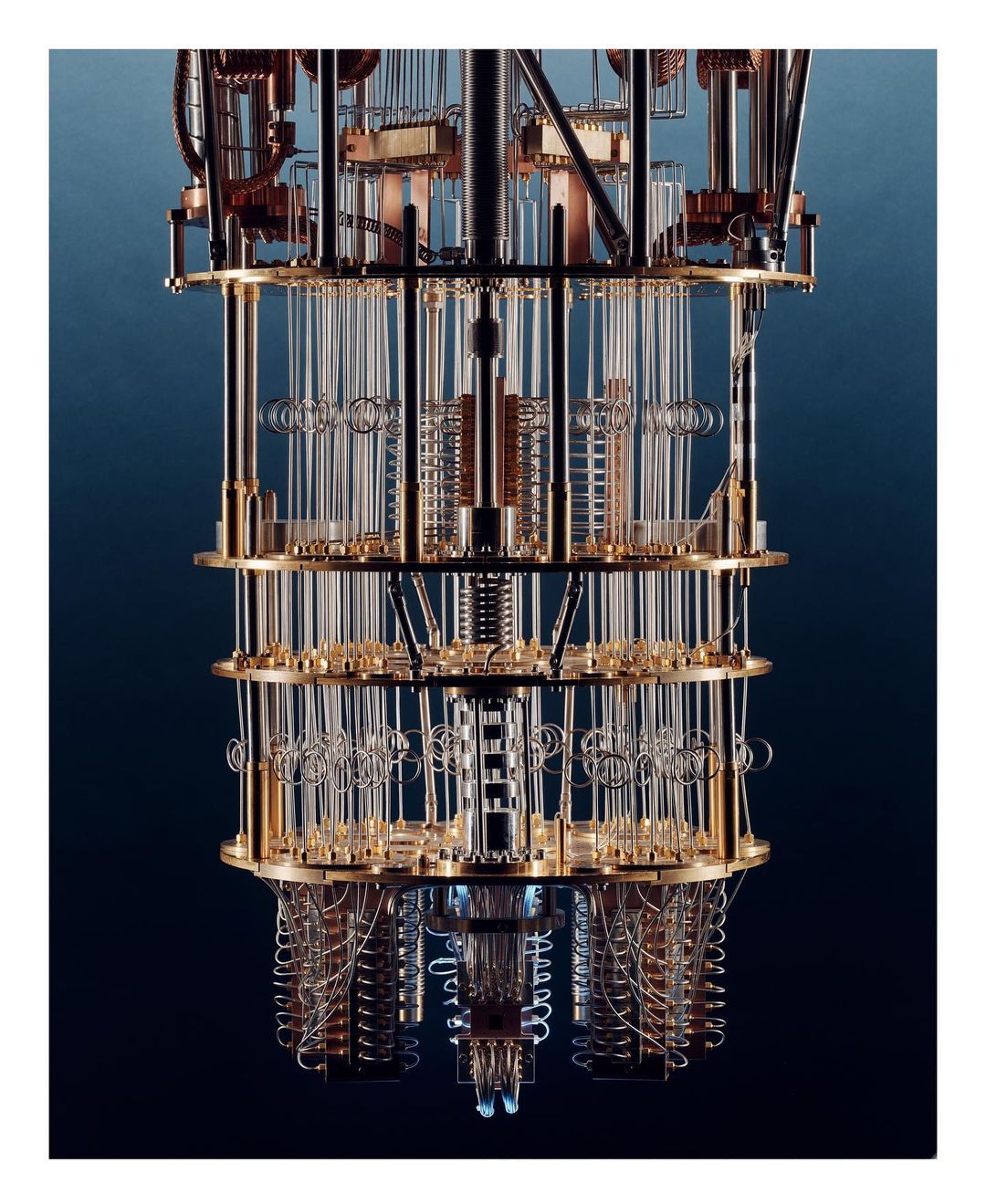 Close-up view of a quantum computing machine with tiered layers of clear tubes, wires, and circular components against a dark blue backdrop.