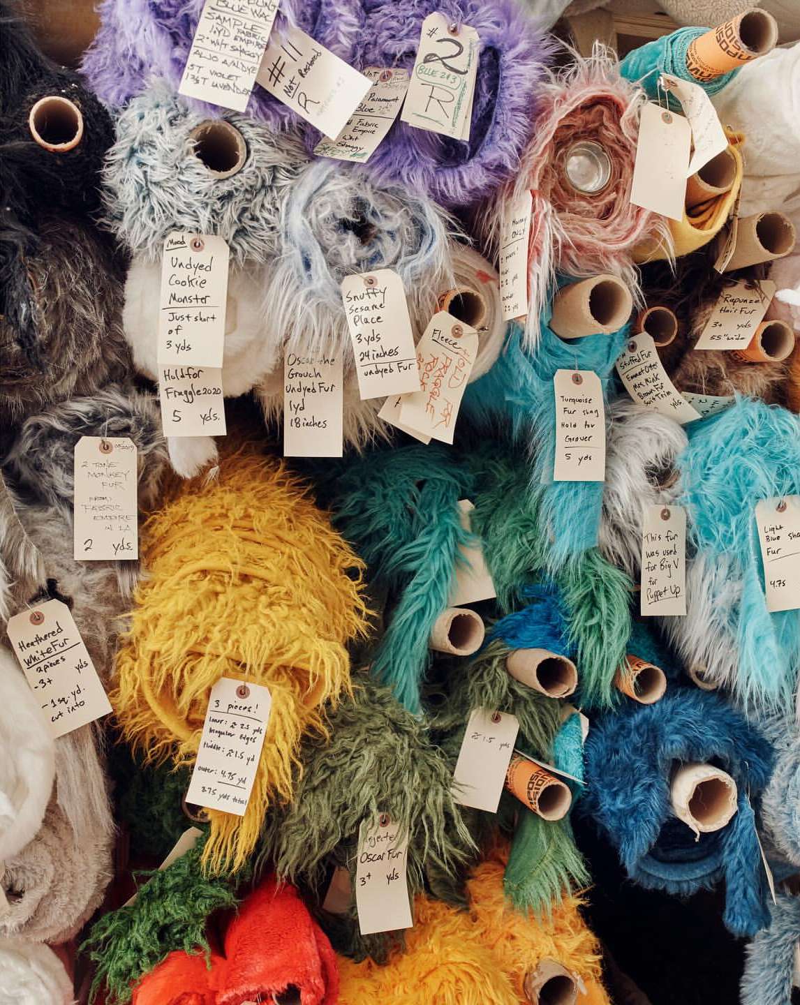 Close-up of many rolls of colorful faux fur fabric with paper tags, stacked together in a fabric store, showing a variety of textures.