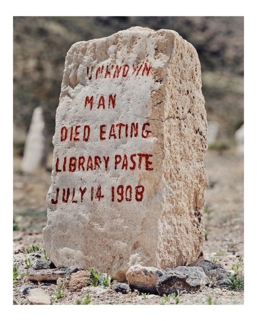 Weathered stone monument with faded red inscription 'Unknown Man Died Eating Library Paste July 14 1908' in an outdoor setting.