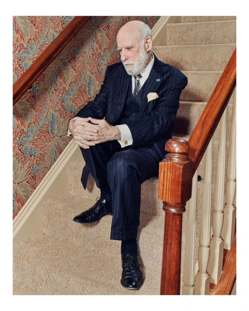 Elderly man in a pinstripe suit sits on carpeted stairs with hands clasped, wearing black leather dress shoes and a tie with a lapel pin.